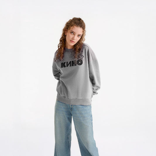 Beautiful young model girl in a photo studio setting wearing official grey KINO band sweatshirt with large КИНО logo on front. From kinoband.com