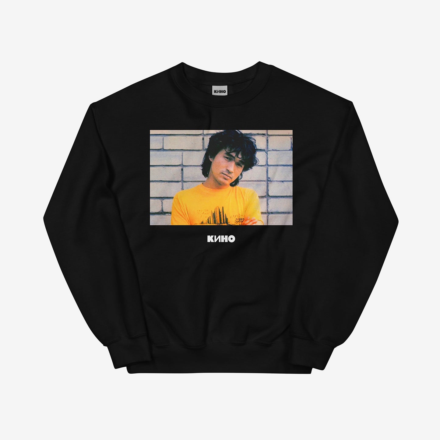 Rare archival photo of Viktor Tsoi in a yellow T-shirt against a white brick wall with a little KINO logo underneath, printed on the front of a heavyweight black unisex sweatshirt with a KINO logo on the back neck side, from www.kinoband.com. Front side.
