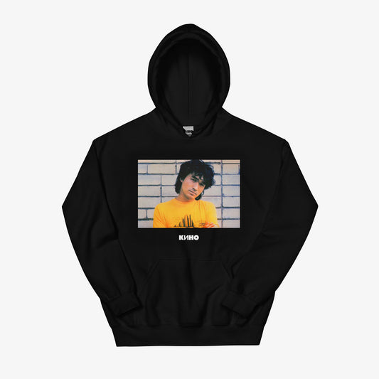 Rare archival photo of Viktor Tsoi in a yellow T-shirt against a white brick wall with a little KINO band logo under the photo, printed on the front of a heavyweight quality black unisex hoodie with a KINO logo on the back neck side, from www.kinoband.com. Front side.