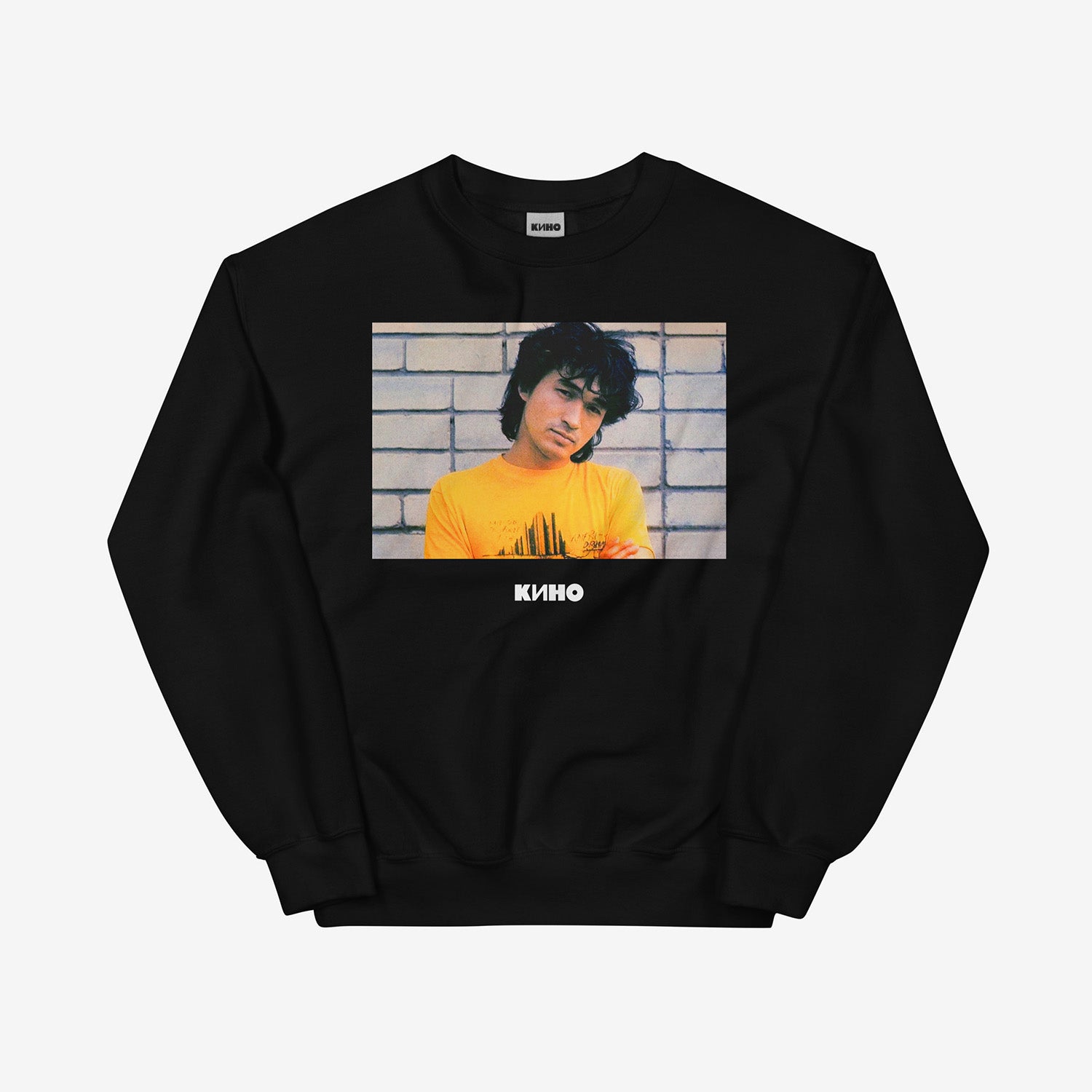 Rare archival photo of Viktor Tsoi in a yellow T-shirt against a white brick wall with a little KINO logo underneath, printed on the front of a heavyweight black unisex sweatshirt with a KINO logo on the back neck side, from www.kinoband.com. Front side.