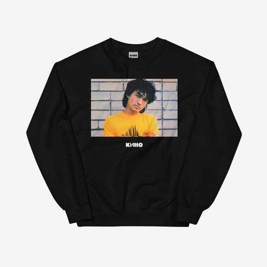 Rare archival photo of Viktor Tsoi in a yellow T-shirt against a white brick wall with a little KINO logo underneath, printed on the front of a heavyweight black unisex sweatshirt with a KINO logo on the back neck side, from www.kinoband.com. Front side.