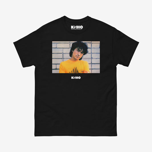 Rare archival photo of Viktor Tsoi in a yellow T-shirt against a white brick wall, printed on the front of a heavyweight black unisex t-shirt with a KINO logo under the photo and on the inside neck label, from www.kinoband.com