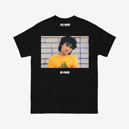 Official KINO band shirt with large print of Viktor Tsoi wearing yellow tee against a white brick wall, with Группа КИНО logo under the photo. www.kinoband.com