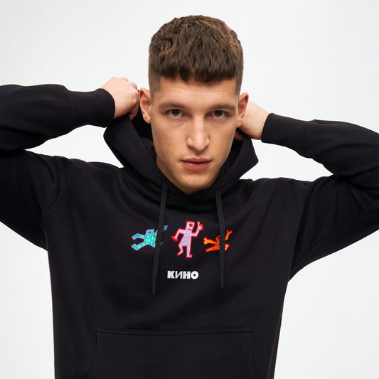 Handsome model boy wearing official black unisex KINO band hoodie featuring an original colorful painting by Viktor Tsoi with a small КИНО logo underneath, with small "Виктор Цой” logo in Cyrillic on the back. www.kinoband.com