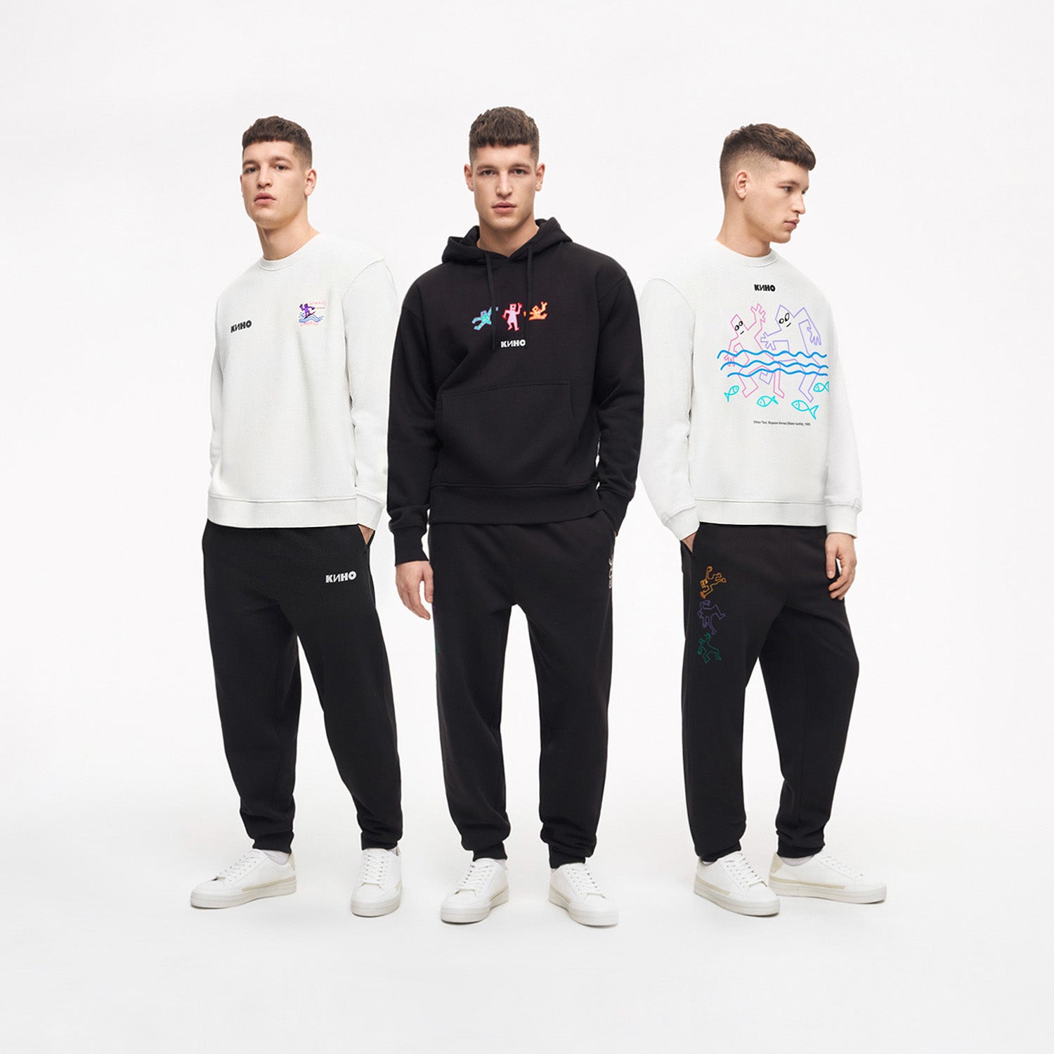 Three handsome model boys standing full length in photo studio, wearing official unisex KINO band hoodie and sweatshirts each featuring original colorful painting by Viktor Tsoi with КИНО logo and small "Виктор Цой” logo in Cyrillic on the back. www.kinoband.com