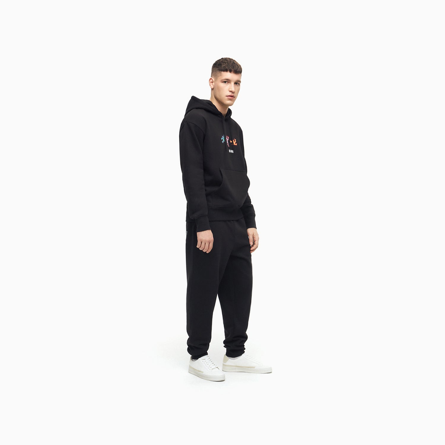 Handsome model boy standing full lenght in photo studio, wearing official black unisex KINO band hoodie featuring original colorful painting by Viktor Tsoi with a small КИНО logo underneath, with small "Виктор Цой” logo in Cyrillic on the back. www.kinoband.com