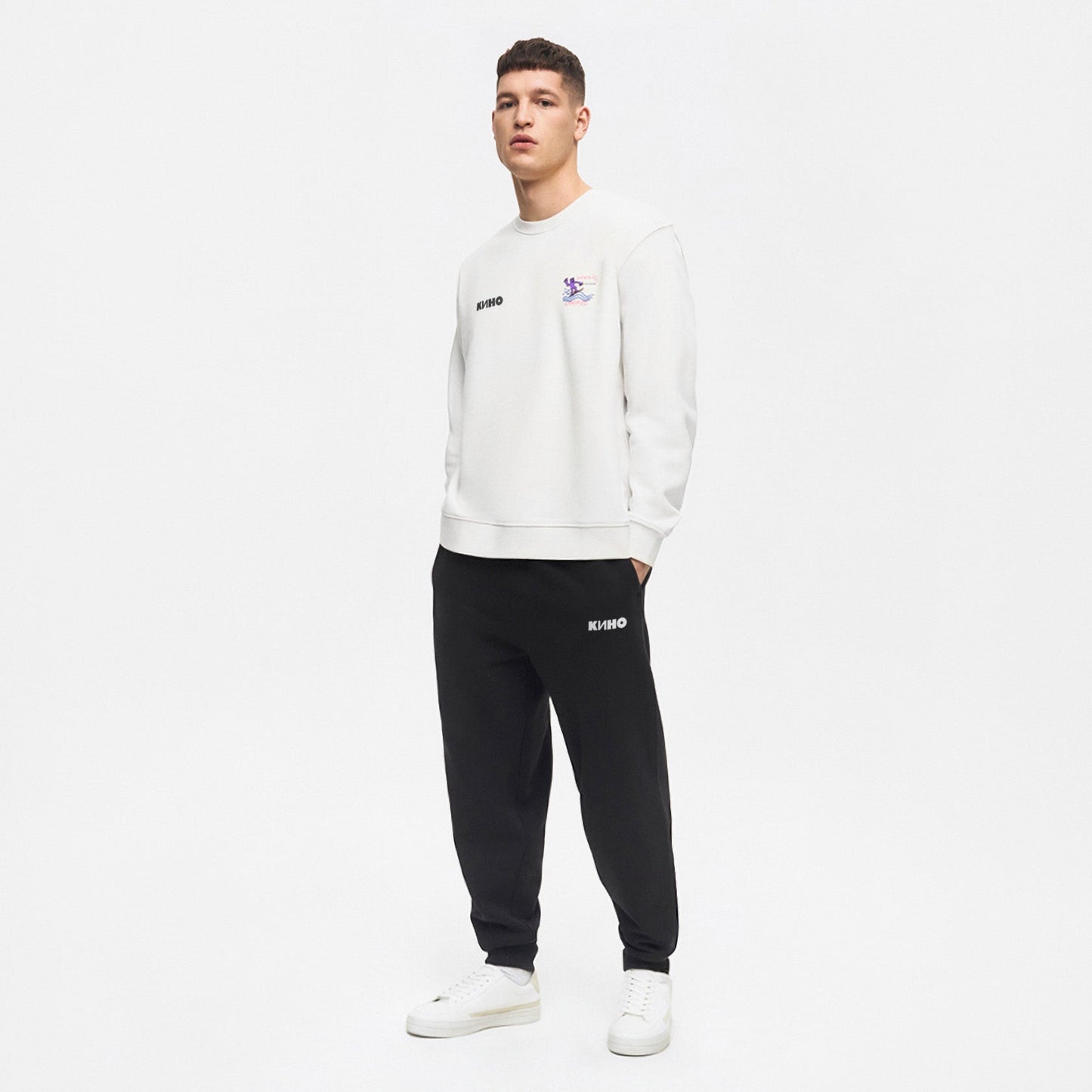 A model boy standing full lenght in a photo studio, wearing white unisex KINO band sweatshirt featuring an original painting by Viktor Tsoi with a small КИНО logo on the chest, with custom necktag logo design  in Cyrillic "Виктор Цой”. Wearing black joggers with KINO band logo. www.kinoband.com